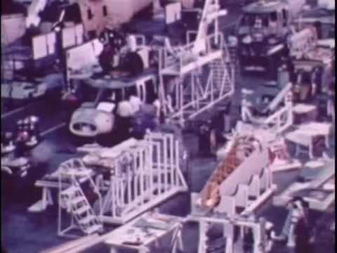 The still from the 1977 Sikorsky S-76 promo film depicts a busy manufacturing environment. Various components and structures related to the S-76 are arranged in an organized manner, with assembly lines and equipment visible. The scene captures a mix of machinery and parts, suggesting an active production process. The overall tone is industrial, reflecting the era's aesthetic and the significance of the Sikorsky brand in aviation. The lack of narration adds a sense of intrigue, leaving viewers to consider the importance of the displayed elements.