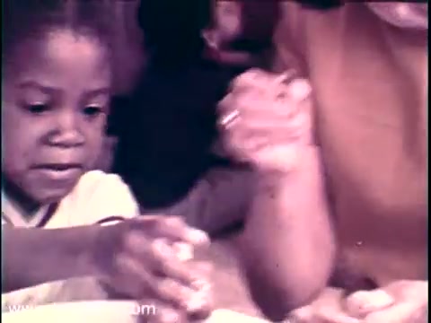 The still captures a moment from the 1968 PSA featuring Lady Bird Johnson, focused on the Head Start program. A young child, intensely engaged, reaches for food while an adult helper guides the experience, emphasizing care and support. The warm color tones highlight a nurturing environment, reinforcing the themes of education and community assistance. This image reflects the program’s mission to support early childhood development.