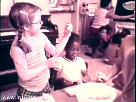 The still from the 1968 PSA 'Head Start,' featuring Lady Bird Johnson, depicts a nurturing classroom scene. A young girl with glasses playfully balances an egg on her head, while a smiling Black boy watches. Children are engaged in various activities, highlighting themes of collaboration and early childhood education. The color palette is soft, typical of 16mm prints from that era. This moment emphasizes the importance of community and learning in the Head Start program. Follow us on Twitter @FromFilm.