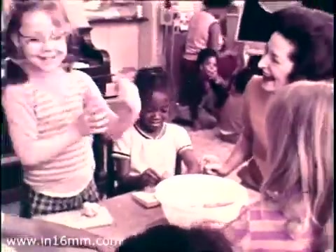 The still from the 1968 PSA 'Head Start' featuring Lady Bird Johnson captures a warm, engaging scene in an early childhood education setting. A young girl in a striped shirt joyfully claps her hands, while another child watches intently. Adult figures, likely educators, interact with the children, creating a nurturing atmosphere. The print's muted colors reflect the era's aesthetic, emphasizing the program's focus on early childhood development and community support.
