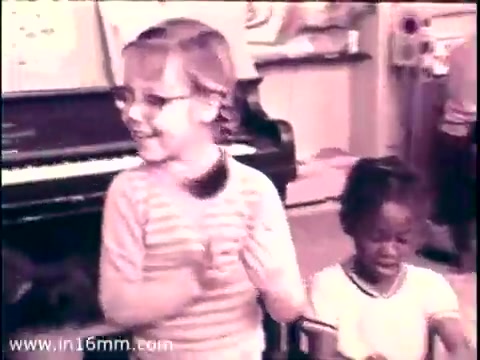 The still features two young children engaged in a classroom setting. One child, wearing glasses and a striped shirt, is smiling and clapping, conveying a sense of joy and participation. Beside her, another child looks focused, seated lower and wearing a simple white shirt. The background suggests an educational environment, highlighted by a piano and various learning materials, underscoring the theme of early childhood education promoted by the Head Start program. The image captures the essence of youthful enthusiasm and learning from the 1968 PSA with Lady Bird Johnson.