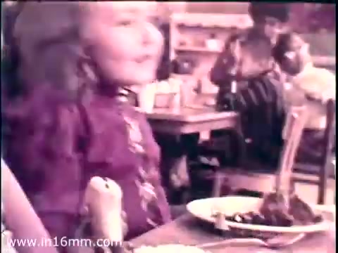 The still from the 1968 PSA 'Head Start' featuring Lady Bird Johnson captures a candid moment in a classroom setting. A young girl, dressed in a colorful outfit, sits at a table with a plate of food in front of her. The atmosphere appears lively, with other children and adults in the background, engaging in activities. This scene reflects the program's emphasis on early childhood education and nutrition. The 16mm film grain adds a nostalgic quality to the image.   Follow us on Twitter @FromFilm.