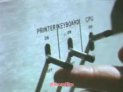 The still from the 'Point Park College PSA - 1969' features a close-up of a hand operating a control panel on a hard copy terminal. The panel includes switches labeled 'PRINTER,' 'KEYBOARD,' and 'CPU,' indicating their operational status. The image captures a moment of technological interface typical of the late 1960s, emphasizing the era’s computing technology. The context suggests a focus on Point Park College's advancements in education and technology during that period.