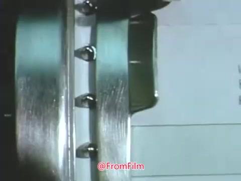 The still features a close-up of a mechanical device resembling a punched card reader or printer, with metal components and a sheet of paper visible. The setting is likely inspired by technology from the late 1960s, reflecting the era's computing methods. This scene relates to Point Park College's focus on advancing education through modern technology.