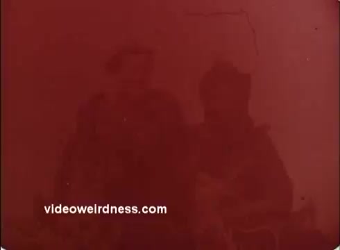 The still appears to be slightly faded or discolored, giving it a red tint. It features two figures, likely individuals associated with the Peace Corps, possibly engaged in a community activity or project typical of their mission in the 1970s. The overall composition suggests a focus on collaboration and service. The image reflects the era's film quality, typical of 16mm footage from that time.