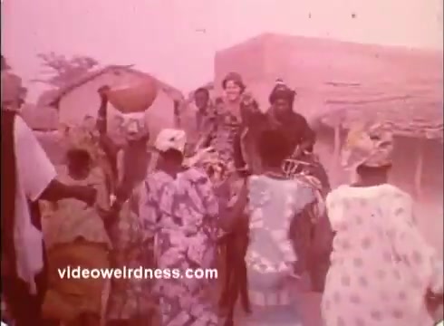 The still captures a joyful gathering in a community setting, likely in an African village, with individuals dressed in colorful traditional garments. A group of people, including women carrying baskets on their heads, are seen celebrating or participating in a communal activity. The scene conveys a sense of cultural vibrancy and togetherness, reflecting the spirit of Peace Corps involvement during the 1970s. Dusty surroundings and simple structures in the background suggest a rural environment.