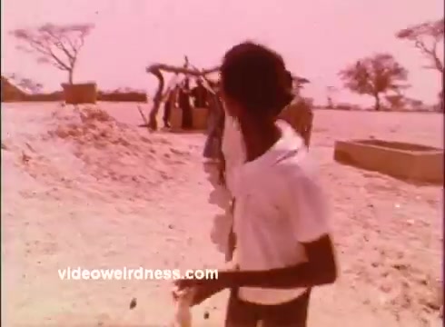 The still from the 1970s Peace Corps PSAs features a young boy in a white shirt, turning away from the camera. He is in a sandy, rural setting with sparse trees in the background. A group of people can be seen in the distance near a structure, possibly engaged in an activity or gathering. The overall color tone has a warm, pinkish hue, indicative of the film style of that era. The scene reflects themes of community and development, common in Peace Corps messaging.