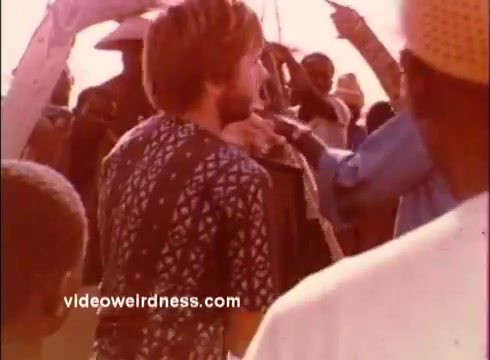 The still features a group of people in a lively scene, likely in an outdoor setting typical of the 1970s. A young man wearing a patterned shirt is the focal point, surrounded by a crowd that appears engaged and animated. The warm tones and vintage aesthetic reflect the era of the Peace Corps PSAs, emphasizing community and cross-cultural interaction.