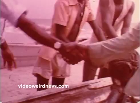 The still depicts a cordial handshake between two individuals, symbolizing partnership or cooperation. The scene is set outdoors, likely by a beach, with several people in the background suggesting a community context. The warm, muted color tones evoke a nostalgic feel typical of the 1970s, aligning with the Peace Corps' mission of fostering connection and support in developing nations.