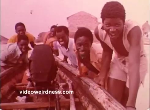 The still depicts a group of joyful young boys leaning toward the camera, showcasing their smiles and lively energy. They are positioned in a wooden boat, suggesting a setting likely related to community or teamwork. The warm, faded colors give it a vintage feel, characteristic of 1970s media. This scene likely serves to promote the positive impact of the Peace Corps in communities during that era.