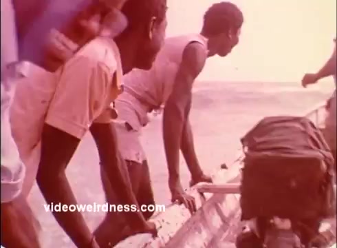 The still features several men interacting with a small boat by the water, likely in a tropical setting. They appear engaged and focused on their task, with personal belongings, such as backpacks, visible. The image conveys a sense of community and collaboration, aligning with the Peace Corps' mission of volunteer service and cultural exchange during the 1970s. The color tone suggests a warm, nostalgic atmosphere typical of that era's media.