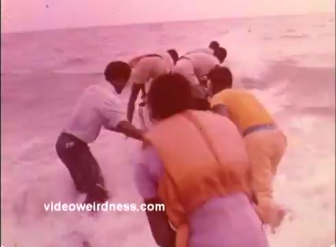 The still depicts a group of individuals wading into the ocean, engaged in an activity that appears to involve teamwork or rescue. The scene has a warm, soft color palette typical of 1970s film, suggesting a focus on collaboration and community service, likely in the context of Peace Corps initiatives promoting aid and support in coastal or underserved regions.
