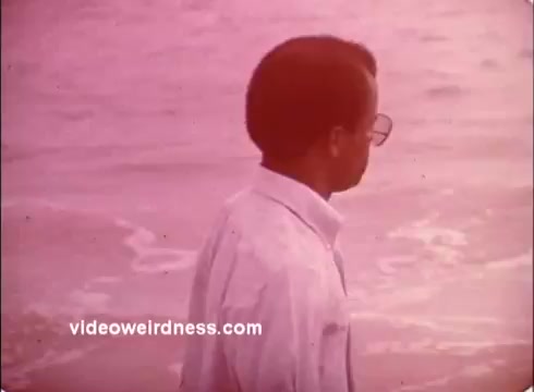 The 16mm still from the 1970s Peace Corps PSAs depicts a man standing by the water's edge, looking contemplative. He wears a white shirt and glasses, and the scene is bathed in a pinkish tint, typical of the era's film style. The image exudes a sense of reflection and calm, aligning with the themes of service and exploration highlighted in Peace Corps messaging.