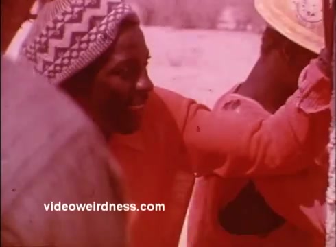 The still from the 1970s Peace Corps PSAs shows a close-up of a smiling individual wearing a patterned headscarf, engaging in a communal activity with others. The image has a warm, reddish tint typical of the era's film style, capturing a sense of camaraderie and connection in a vivid, nostalgic manner. The context suggests themes of community involvement and the spirit of the Peace Corps mission.
