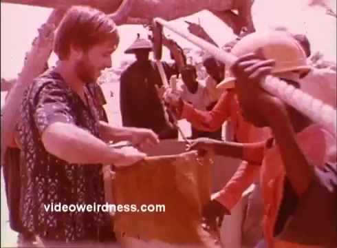 The still depicts a scene from a 1970s Peace Corps public service announcement. A Western volunteer, dressed in a patterned shirt, engages with local workers, focused on a task involving fabric or material. The setting appears to be an outdoor work site, with several individuals in the background, some wearing helmets, emphasizing community collaboration. The overall tone suggests a message of international cooperation and volunteerism.