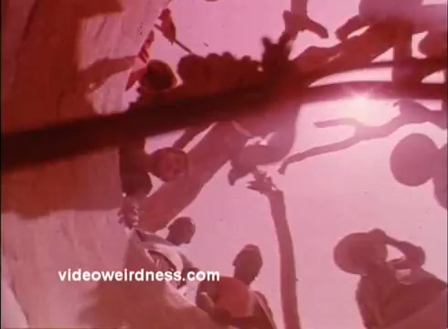 The 16mm still from the 1970s Peace Corps PSAs depicts a group of people engaged in a communal activity, seen from a low angle. The image has a warm, pinkish hue, and the scene appears lively, reflecting themes of collaboration and community. The perspective emphasizes the inclusivity and participatory spirit of the Peace Corps mission during that era.