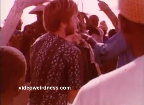 The still depicts a scene from a 1970s Peace Corps PSA. In the foreground, a man with a shaggy hairstyle is wearing a patterned shirt, representing a typical volunteer. Surrounding him are several individuals, likely locals, engaged in a lively interaction, indicating cultural exchange. The warm color tones create an inviting atmosphere, typical of the era's film style. The image captures the spirit of community and the mission of the Peace Corps during this time.