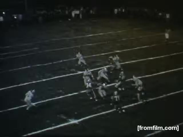 The still captures a high school football scene at night, featuring players on the field in action. Some players are positioned to receive the ball, while others appear ready to defend. The field is marked with white lines, and a small crowd is visible in the background, enhancing the atmosphere of a local football game. The image is in monochrome, typical of the 16mm film format.