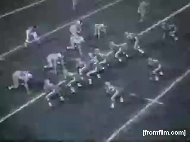 The still features a high school football game captured in black and white. Players are in a formation on the field, with some in huddle positions and others lined up ready for a play. The scene conveys a dynamic moment typical of high school football, highlighting the strategy and teamwork involved in the game. The setting appears to be a grassy football field, with yard lines clearly marked.