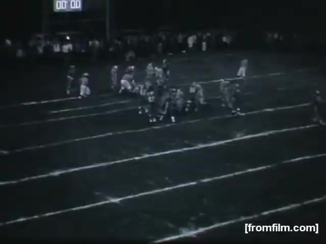 The still depicts a high school football game under night lighting, showcasing players positioned on the field. The scoreboard displays a time of 00:00, indicating the end of a game or quarter. The players are in action, with the field marked by clear yard lines suggesting a competitive match. A crowd can be seen in the background, adding to the atmosphere of the event. The overall tone is nostalgic, reflecting the spirit of high school sports.