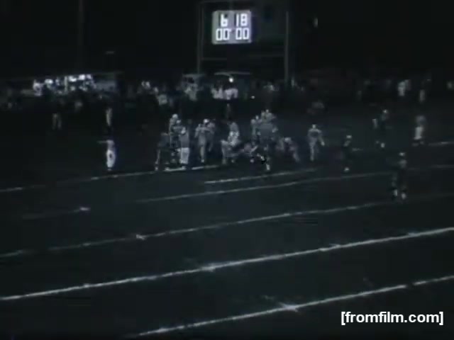The still captures a nighttime high school football game, showing players huddled together on the field. A scoreboard in the background displays '6:18' and '00:00,' indicating time left in the game or quarter. The scene is illuminated by stadium lights, with a crowd visible in the background, adding to the atmosphere of a competitive sporting event. The focus is on the players, highlighting the teamwork and strategy typical of high school football.