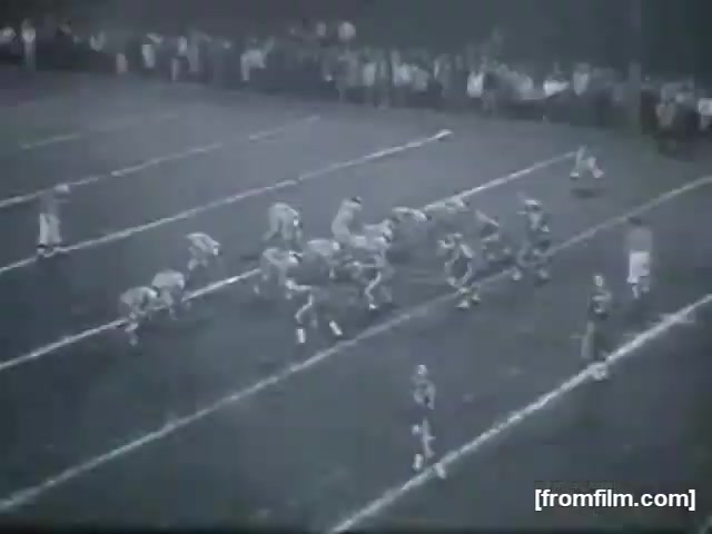 The still captures a high school football game, showing players from two teams lined up on the field. The scene depicts a moment before the snap, with one team in a huddle and the opposing team ready for defense. Spectators can be seen in the background, suggesting a lively atmosphere. The film has a vintage quality typical of 16mm footage, emphasizing the historical context of high school football.