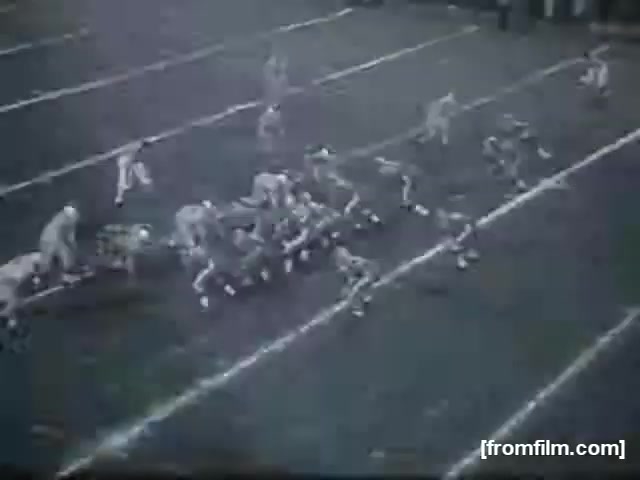 The still features a high school football game, capturing a moment during play. Players in uniforms are positioned on the field, with one team lined up for a play. The scene suggests a competitive atmosphere typical of high school football, with marked yard lines and a well-defined playing field. The image has a vintage quality, indicative of 16mm film.