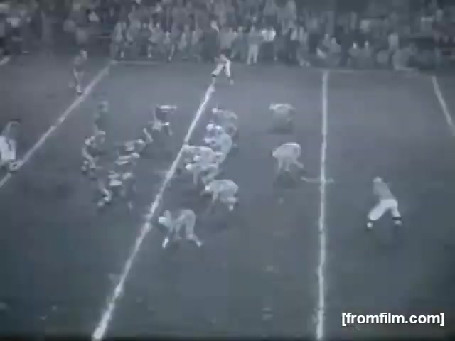 The still presents a high school football game featuring two teams lined up on the field. One team is positioned in a huddle, ready to execute a play, while the other team is aligned defensively. The field is marked with yard lines, and a crowd can be seen in the background, capturing the atmosphere of a live sports event. The monochromatic quality of the film emphasizes the era's vintage aesthetic.