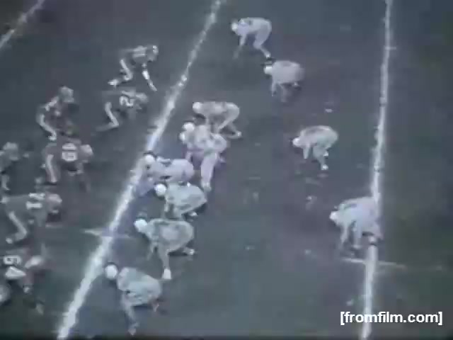 The still captures a high school football game, showing two teams lined up on the field. The players, in different colored uniforms, are positioned for a play, with one team in a defensive stance and the other preparing to execute a play. The field is marked clearly with yard lines, typical for football games. The black-and-white film reflects the classic style of 16mm footage.