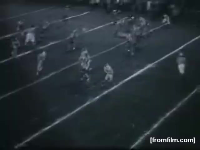 The still captures a high school football game, showing players in action on the field. The athletes are spread across the turf, and the game appears to be in progress. The lighting is dim, typical of older footage, and the focus is on the players as they engage in a play. Lines mark the field, highlighting the gridiron layout. The overall atmosphere conveys the energy and intensity of high school football.