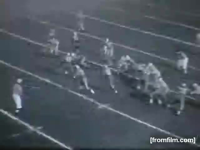 The still from 'High School Football (Reel 3)' depicts a moment during a football play. Players in uniforms are scattered across a marked field, with one player poised to pass the ball. The atmosphere suggests excitement and action typical of high school football games, featuring players in distinct positions, ready for the next move. The black-and-white format gives it a vintage feel.