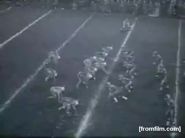 The still from 'High School Football (Reel 3)' depicts a high school football game featuring two teams positioned on the field. The players are in a huddle formation, preparing for the next play. The field lines are clearly marked, and there is a visible crowd in the background, indicating an active game atmosphere. The image has a vintage quality typical of 16mm film, suggesting it captures a moment from a past era of high school sports.
