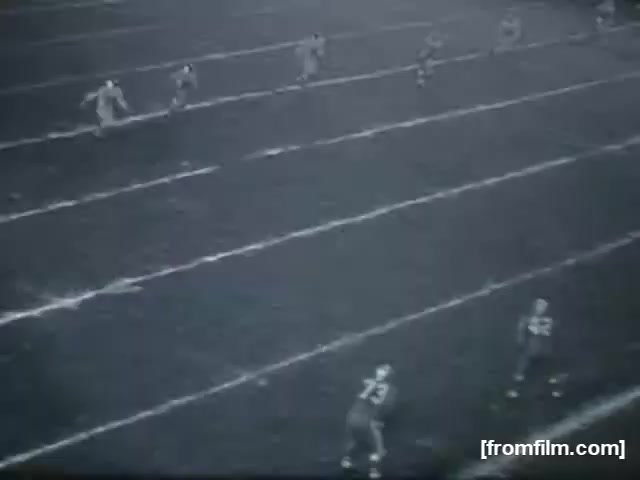 The still depicts a high school football scene captured on 16mm film. Several players are positioned on the field, ready for a kickoff. The players are in contrasting uniforms, indicating two different teams, while the field shows clear markings. The black-and-white imagery gives it a vintage feel, typical of mid-20th century sports footage. The atmosphere appears focused on the impending action of the game.