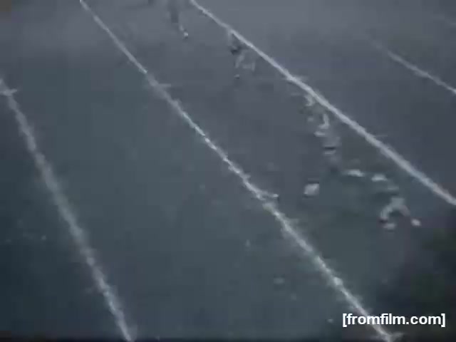 The still from 'high school football (reel 3) [FF0006]' shows a scene on a football field. The image captures players running on a grass field marked with white yard lines. The focus appears to be on the action, highlighting the movement of the athletes in an outdoor sports setting. The atmosphere suggests a live game, featuring the youthful energy typical of high school football.