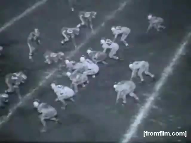 The still captures a high school football scene, showcasing two teams lined up on the field. Players are positioned in various stances, with defenders ready to engage and the offense set for a play. The field markings are visible, and the monochromatic film adds a nostalgic, vintage feel to the atmosphere of the game.