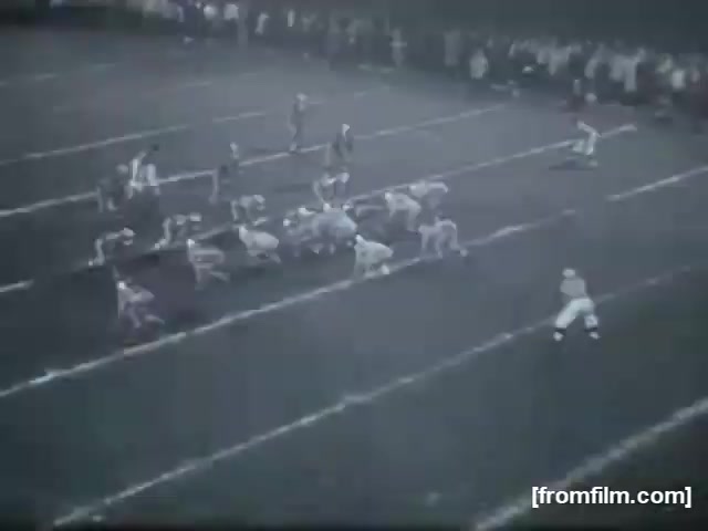 The still features a high school football game with players lined up on the field. The scene captures a moment before the snap, showcasing a formation with a focus on the players' positioning and the field markings. Spectators are visible in the background, indicating an engaged crowd watching the game. The overall atmosphere reflects the competitive spirit of high school football.
