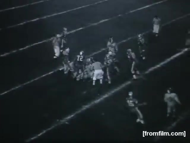 The still features a high school football game, capturing a moment with players from both teams engaged in an active play. The scene is set on a gridiron field, marked by white yard lines, and players are wearing uniforms, with one player prominently displaying the number 72. The atmosphere suggests a tense moment of competition, with players focused on the ongoing action. The image is presented in a monochrome style typical of 16mm film.
