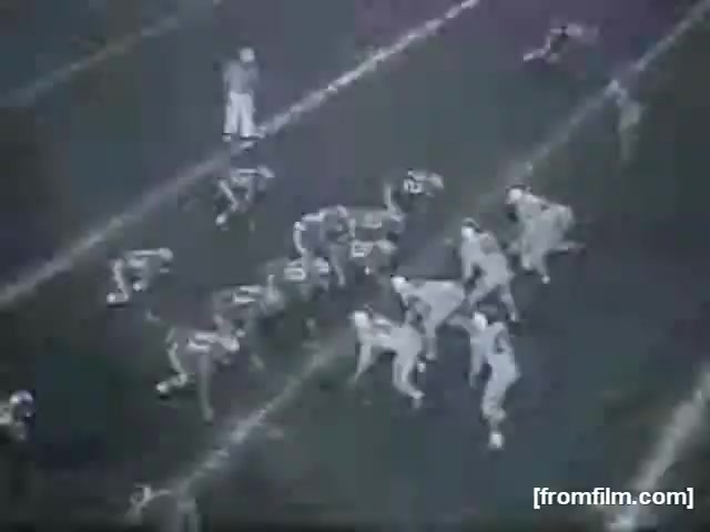 The still features a high school football game, with two teams positioned on the field. Players are lined up in formation, preparing for the play. The scene captures the intensity of the moment, showcasing the athleticism and strategy inherent in high school football. The image is in monochrome, typical of 16mm film, adding a nostalgic quality to the action.