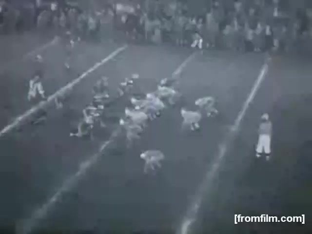 The still captures a high school football game, showing players lined up on the field. The scene features a clear view of the formation, with several players in action, while a crowd can be seen in the background, suggesting a lively atmosphere. The monochrome footage adds a vintage feel to the moment, typical of 16mm film.
