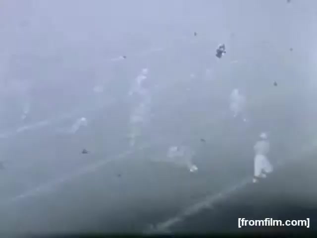 The still from 'high school football (reel 3) [FF0006]' depicts a foggy scene on a football field. Players in light-colored uniforms are visible, moving across the field amid a hazy atmosphere. The blurred background suggests poor visibility, typical of an overcast or misty day during a game. The overall mood is somewhat enigmatic due to the lack of clarity.