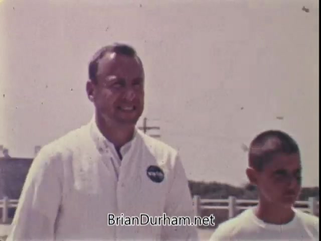 The still features Captain Jim Lovell, dressed in a white NASA jacket, walking alongside a young boy who has a shaved head. Lovell wears a smile, conveying enthusiasm and approachability. The background suggests an outdoor setting, perhaps during a physical fitness event, highlighting the theme of health and fitness advocated by the President's Council on Physical Fitness. The overall tone is inspirational, emphasizing the idea that anyone, regardless of background, can achieve great things.