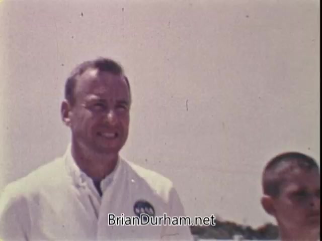 The still features Captain Jim Lovell, an astronaut, in a light-colored jacket with a NASA logo, standing alongside a young boy. Lovell's expression conveys encouragement and enthusiasm, emphasizing the message about physical fitness and ambition. The background appears bright and slightly blurred, typical of 16mm film, enhancing the nostalgic feel of the scene. The overall tone reflects inspiration and the idea that anyone can achieve their dreams with effort, as highlighted in his message about the presidential physical fitness award for youth.