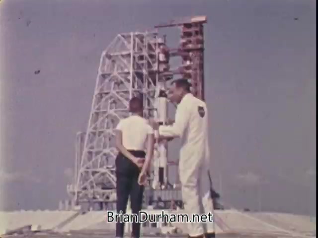 The still features astronaut Jim Lovell in a white NASA jumpsuit, engaging with a young boy dressed in a black outfit and a white shirt. They stand in front of a large rocket launch pad structure against a clear sky. Lovell appears to be explaining something, likely related to physical fitness or aspirations of becoming an astronaut, reflecting the message of the President's Council on Physical Fitness. The scene captures an inspiring moment, promoting the idea that dreams are achievable through fitness and determination.