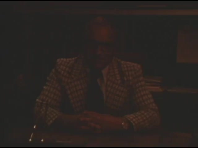 The still from the 1976 political commercial features Stephen Dubetz, an engineer, speaking against Ohio's Issue 6, which aimed to ban the construction of future nuclear power plants. He emphasizes the importance of nuclear energy for the state's progress and warns that passing the issue would hinder development. The setting appears formal, likely in an office, with Dubetz dressed in a distinctive plaid suit, enhancing his authoritative stance.