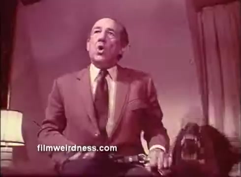 The still features a man seated, passionately expressing himself—possibly mid-sentence—while gesturing. He's dressed in a brown suit with a tie, and there's a dimly lit room behind him, suggesting an intimate or tense setting. A shadowy figure, likely a dog, is beside him, adding to the atmosphere. The image reflects a dramatic moment typical of a thriller like 'The Mackintosh Man.'