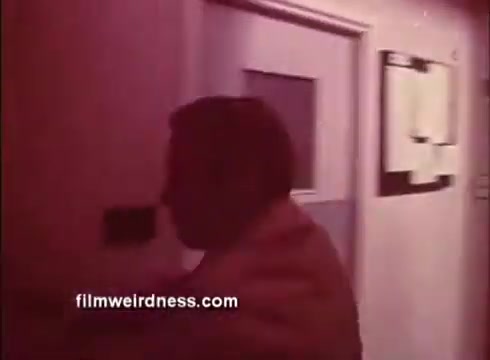 The still from the trailer of 'The Mackintosh Man' features a shadowy figure, likely Paul Newman, moving through a corridor. The lighting creates a dramatic effect, emphasizing the sense of mystery and tension. The environment appears to be a secure or institutional setting, hinting at the film's themes of espionage and intrigue. The overall color tone gives it a vintage feel, typical of 16mm film.