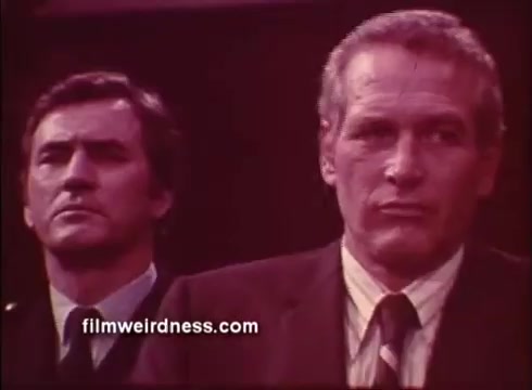 The still features two men, with Paul Newman prominently in the foreground, wearing a suit and a serious expression. The man behind him appears to be in a uniform, suggesting a tense or dramatic situation. The color palette suggests a vintage feel, characteristic of the film's era, likely emphasizing themes of intrigue and suspense from the trailer of 'The Mackintosh Man.'