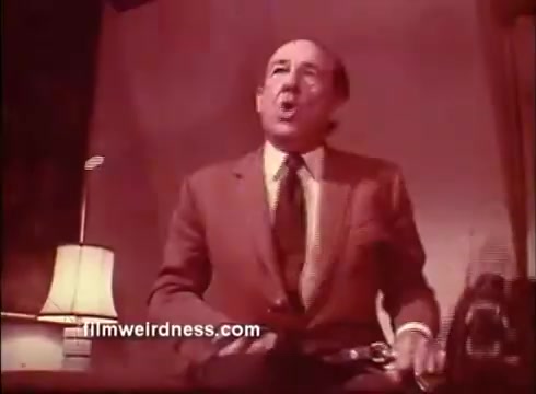 The still from the trailer of 'The Mackintosh Man' features a man in a suit, seated and animatedly speaking. The setting appears somewhat formal, with a lamp providing soft lighting. The expression on his face suggests urgency or intensity, fitting for a suspenseful narrative. The overall tone implies a dramatic scene likely tied to the film's espionage themes.