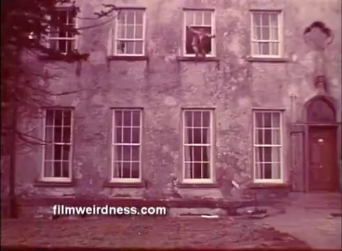 The still depicts a weathered building with several large windows, one of which shows a figure, suggesting a dramatic escape or confrontation. The exterior is marked by age, reflecting a tense and gritty atmosphere typical of thrillers. This scene likely contributes to the mysterious and suspenseful tone of 'The Mackintosh Man.'