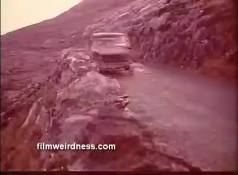 The still from 'The Mackintosh Man' trailer features a vehicle navigating a rugged, rocky road, set against a backdrop of a barren, mountainous landscape. The muted color tones and dramatic scenery evoke a sense of isolation and tension, aligning with the film's themes of intrigue and espionage.